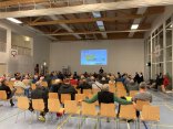 Bürgerversammlung. Menschen sitzen auf Holzstühlen in einer Sporthalle und sehen sich eine Präsentation auf einer Leinwand an